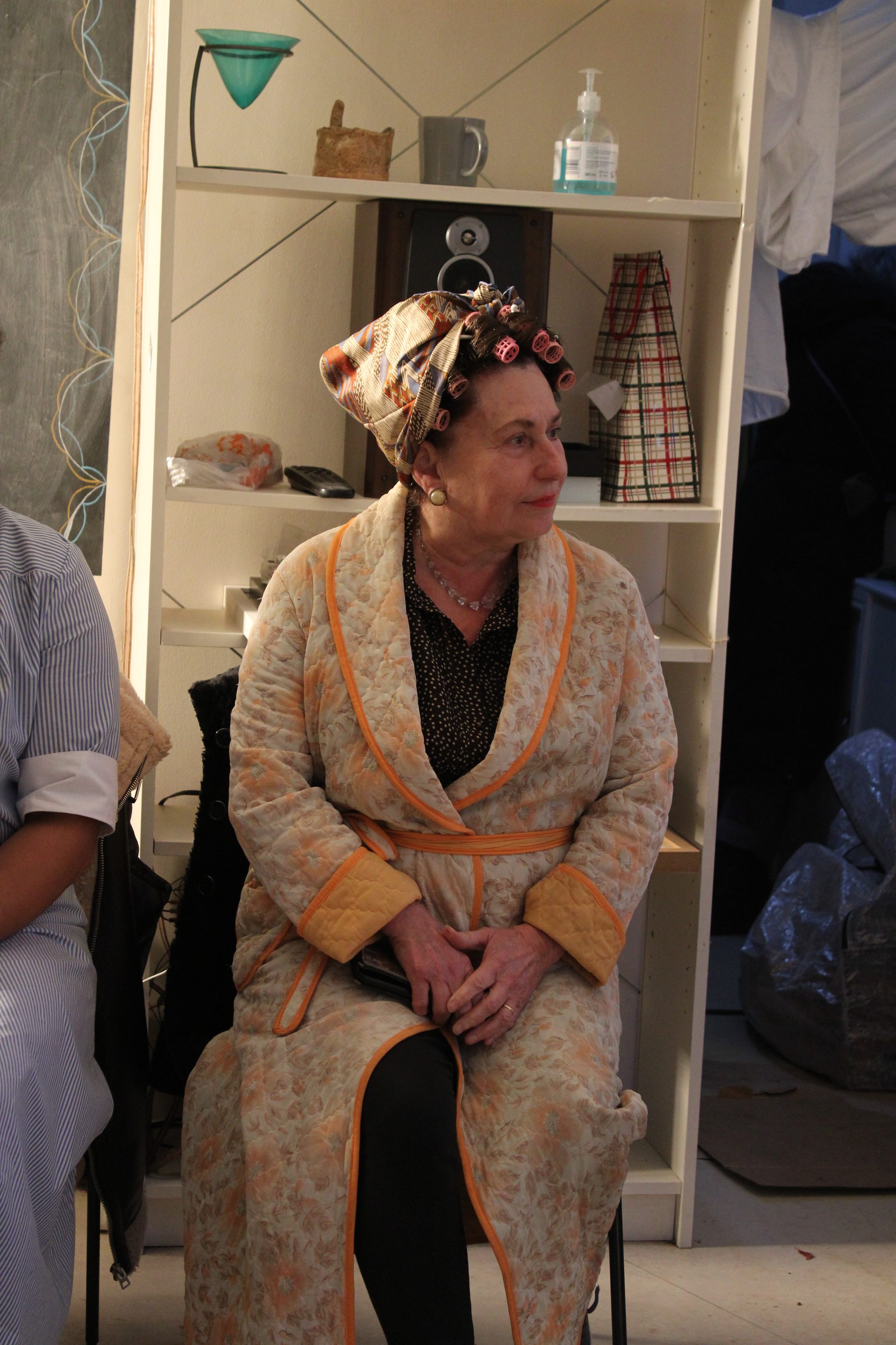 Production photograph: a woman in period costume and headscarf seated in the green room between takes.