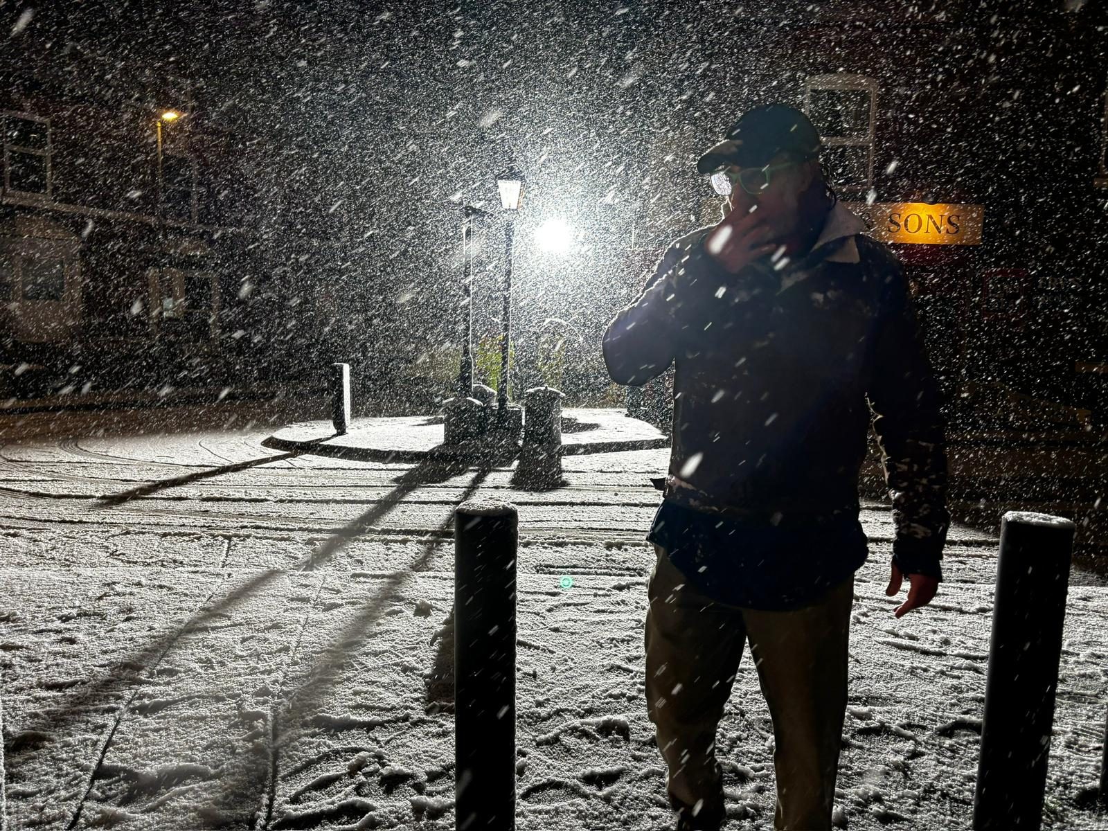 Production photograph: a man smoking under a street lamp on the dressed Butts Road set, snow falling hard through the light.