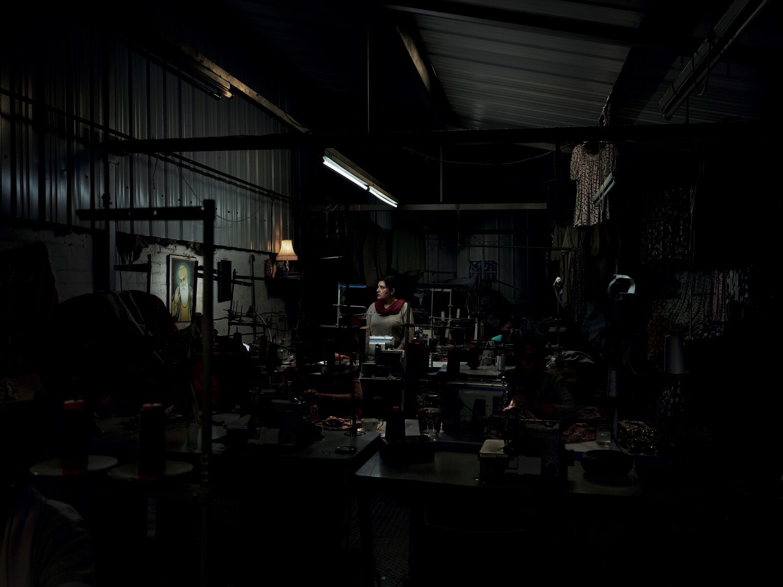 Seamstress, from The Exiles: a woman alone in a darkened sewing factory under a single fluorescent strip, an image of Guru Nanak in the foreground.