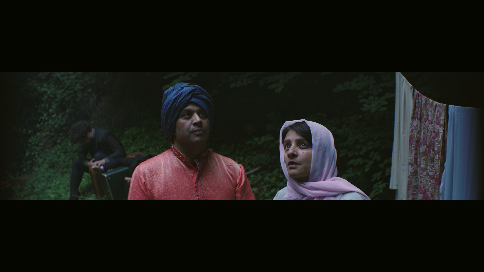 Indi film still: a couple in a garden at night, the man in a blue turban and red kurta, the woman in a lilac dupatta, a figure watching from the shadows behind.