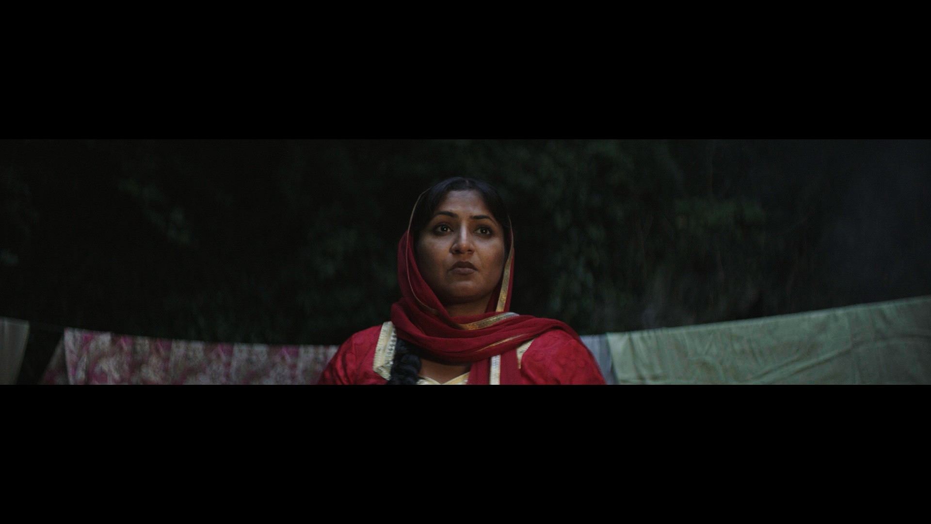 Indi film still: close-up of the mother in a red dupatta, washing line behind her in the dark.