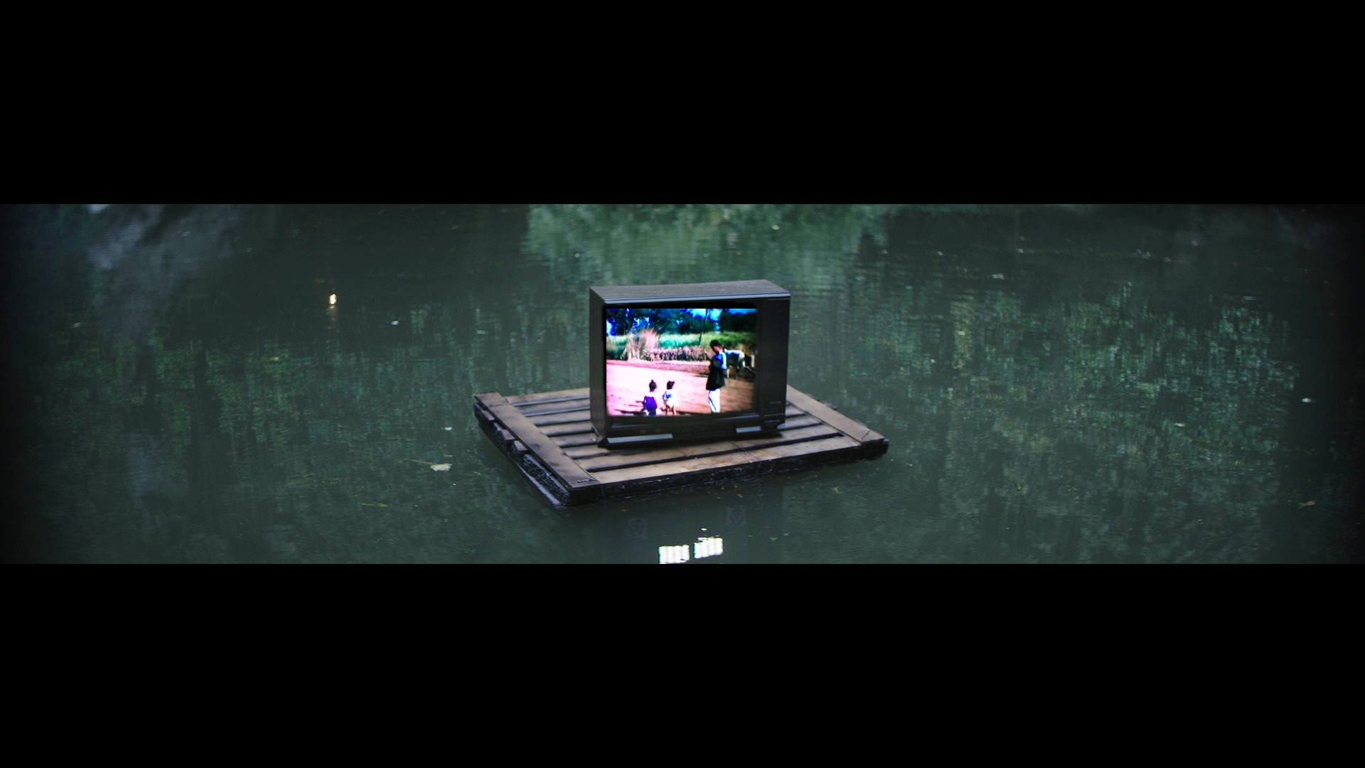Indi film still: a CRT television on a wooden pallet floating on green canal water, a Bollywood film playing on the screen.