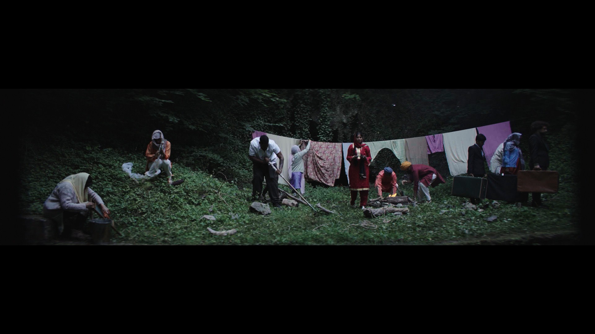 Indi film still: a woodland camp at night, figures gathered around a fire, washing strung between trees, suitcases on the ground.