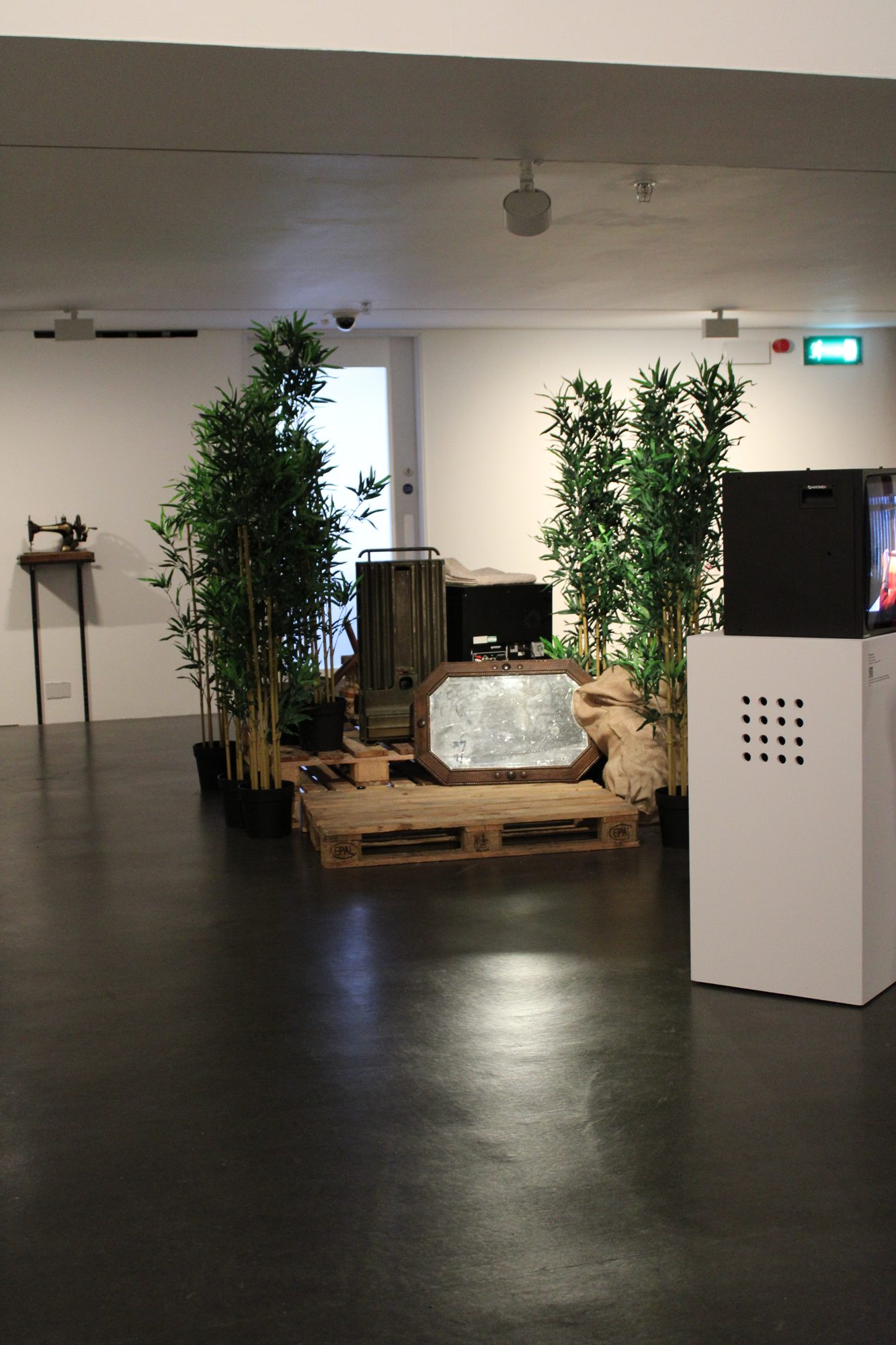 Paths You Walk installation at The New Art Gallery Walsall: a vintage mirror, ammunition box, and burlap arranged on wooden pallets among bamboo plants.