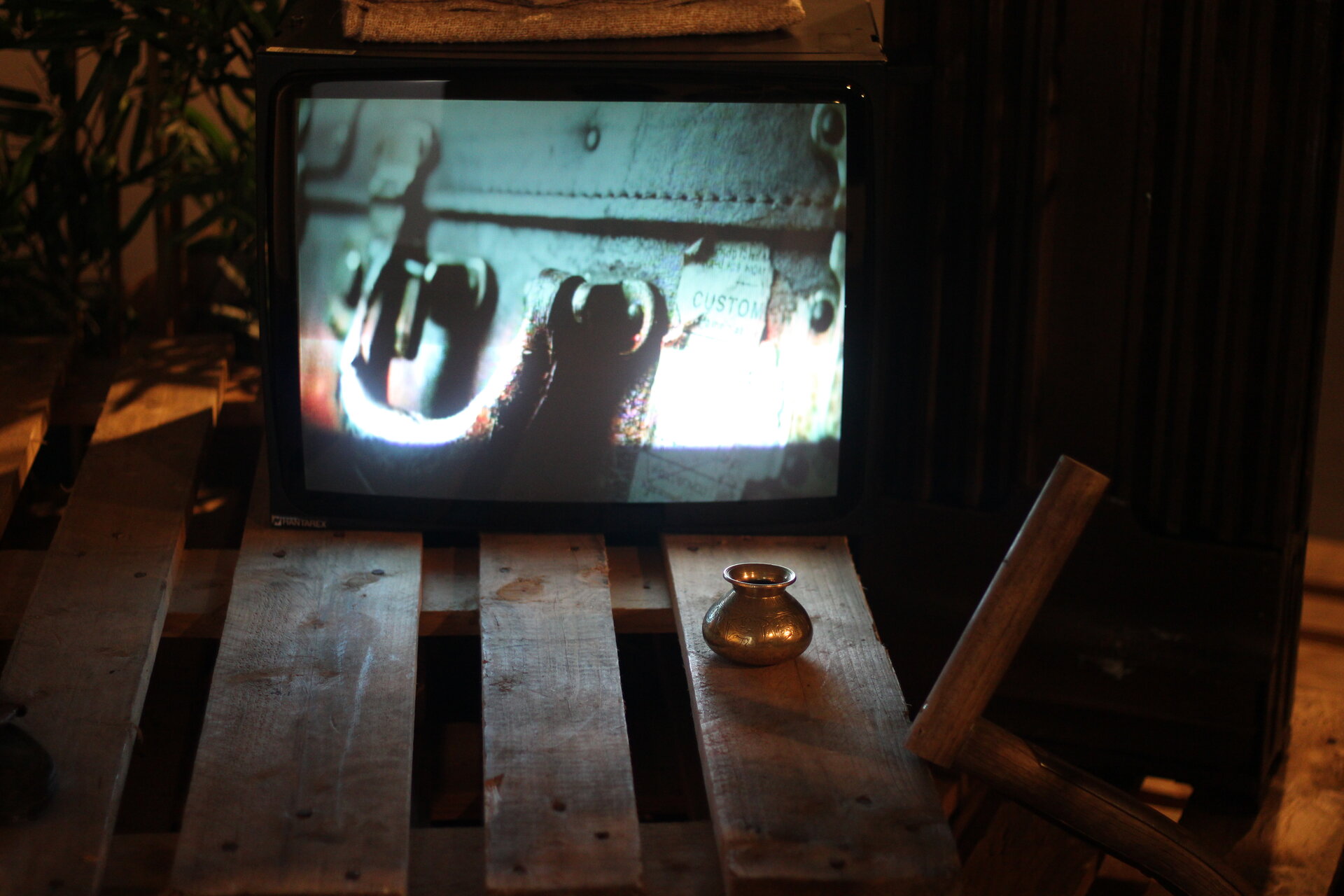 Three Cantos installation: CRT television on a wooden pallet showing film footage, brass lota beside it.
