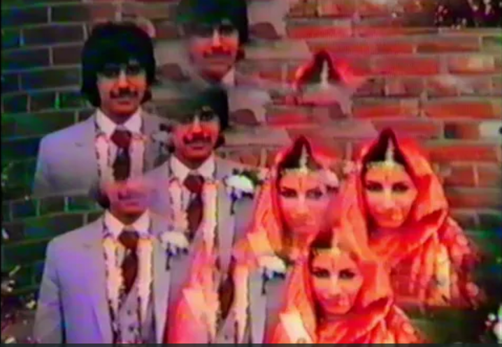 Degraded colour home-movie frame: a Sikh wedding group photographed against a brick wall.